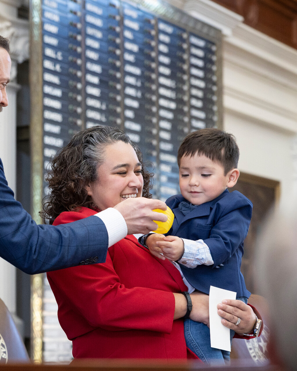 Denise smiling as her son Zeke plays with a yellow toy on the Texas House floor.