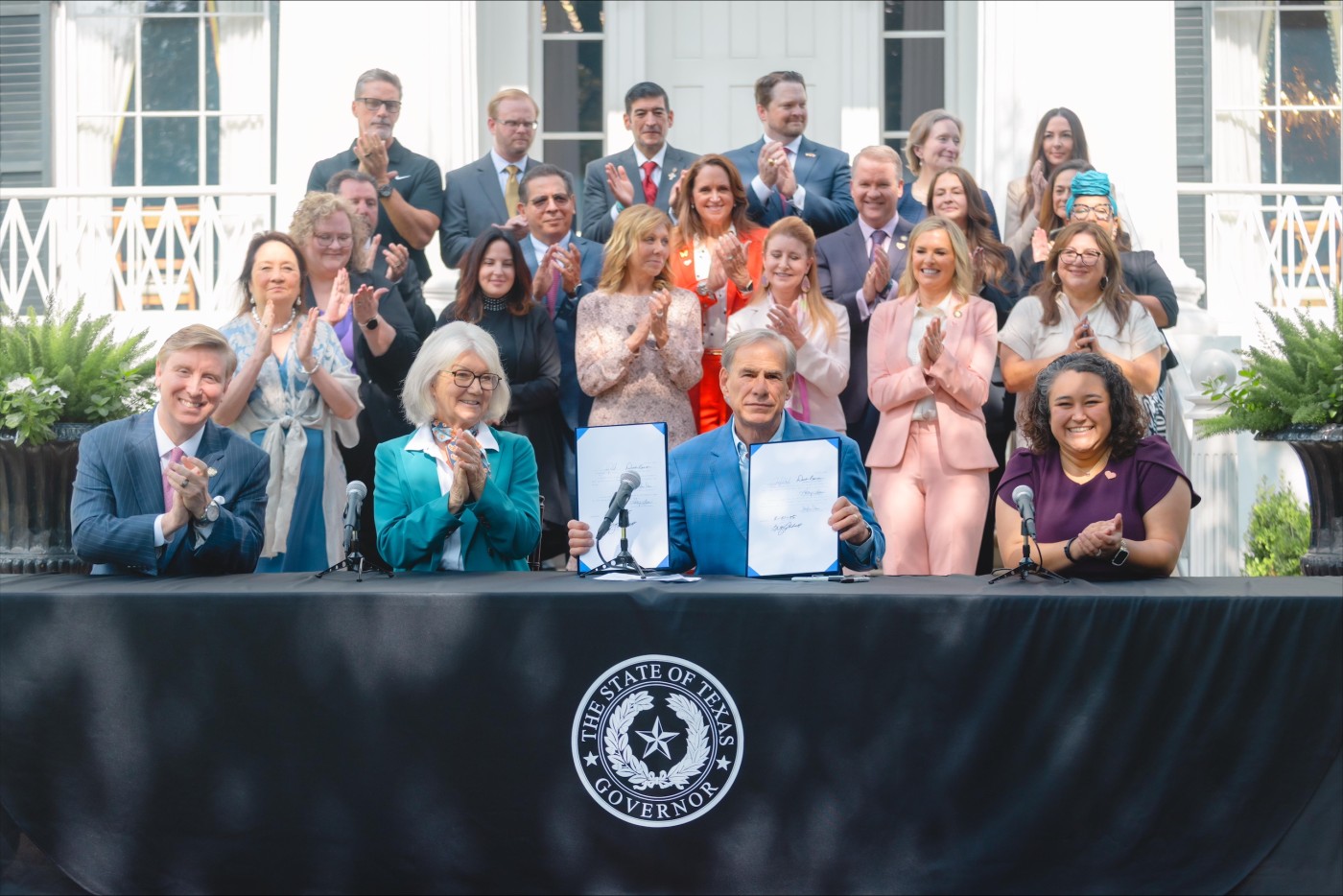 Governor Greg Abbott holds the signed HB 2306 at the Governor's Mansion with Rep. Denise Villalobos seated nearby.